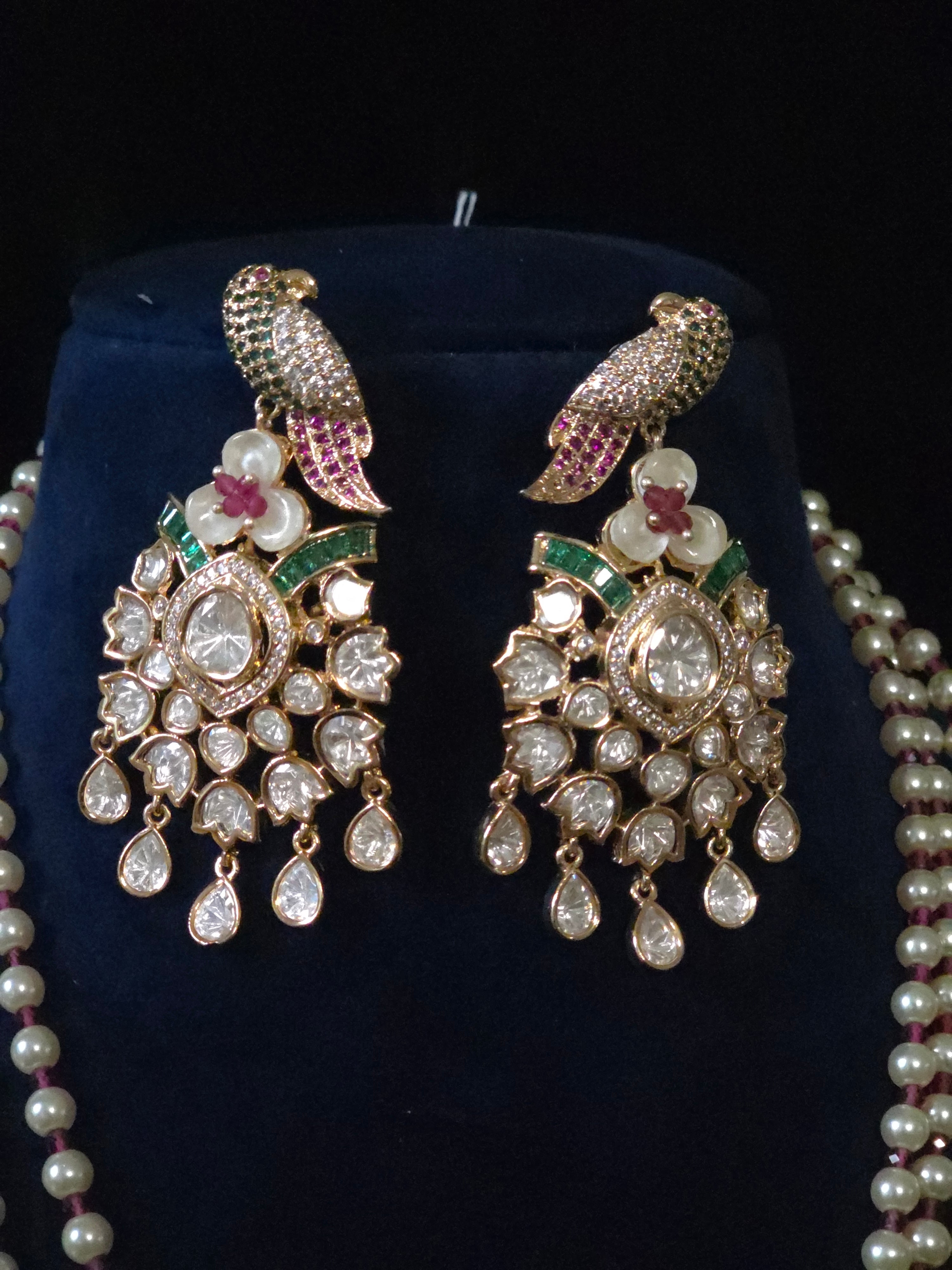 Gold-plated traditional Indian parrot earrings with pearl floral details, pink stone accents, and dangling clear stones, displayed on a dark blue velvet stand alongside a pearl necklace.