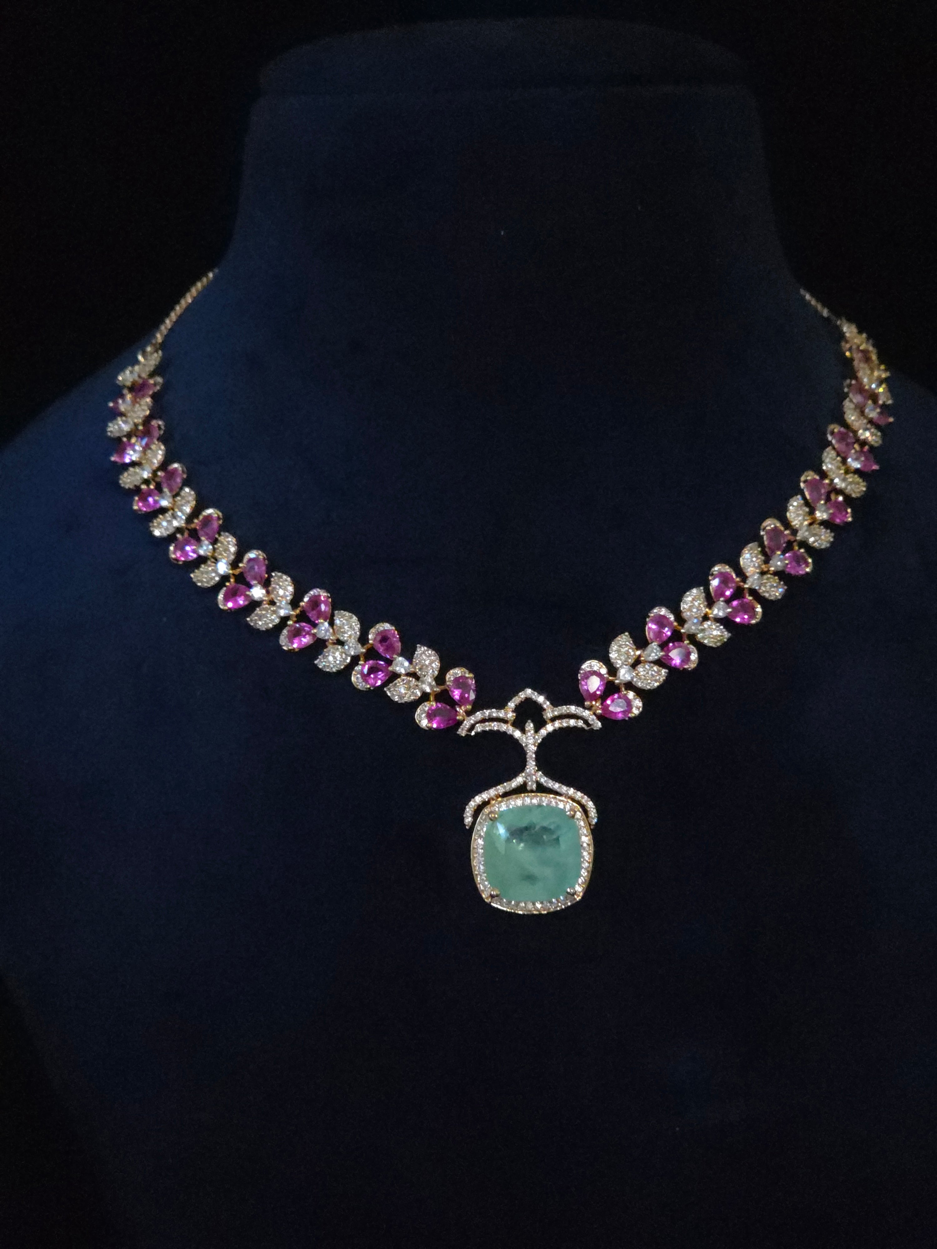 An ornate gold necklace displayed on a black velvet jewelry bust. The necklace features a large central light green rectangular pendant surrounded by small clear stones, with rows of pink gemstones and clear stones forming leaf-like patterns along the chain.