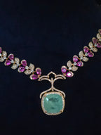 An ornate gold necklace featuring a large cushion-cut green gemstone pendant, surrounded by small clear stones and accented with pink gemstones along the chain, displayed on a dark background.