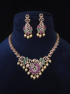 Gold-toned Indian-style jewelry set featuring a necklace and matching earrings with pink gemstones, green stones, and pearl-like bead drops, displayed on a dark blue velvet bust against a black background.