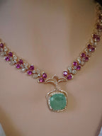 A gold-plated necklace displayed on a beige jewelry bust, featuring a large central green cushion-cut stone surrounded by small clear stones, with rows of pink gemstones along the chain.
