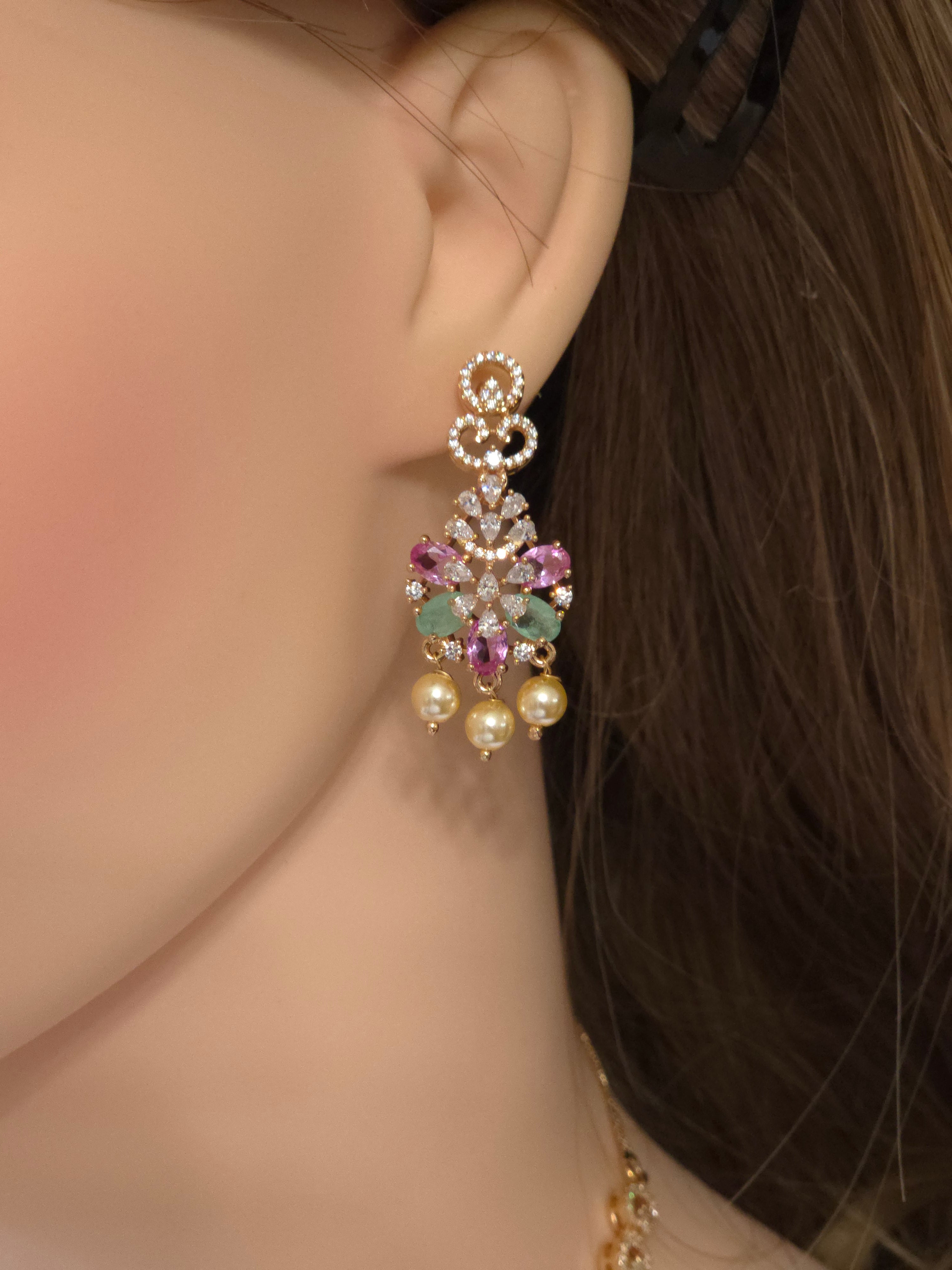 A close-up of a model's ear wearing a dangling chandelier earring. The earring features a gold-toned filigree top, pink and green gemstone accents, and three dangling cream-colored pearl-like beads.