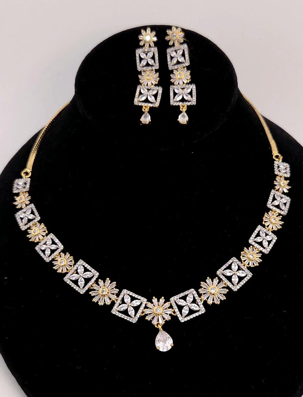 Gold and silver-tone jewelry set featuring a necklace and matching earrings displayed on a black velvet bust. The necklace has repeating geometric and floral motifs with a teardrop pendant, and the matching earrings are shown above the bust against a light gray background.