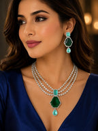 A woman wearing a luxurious jewelry set featuring a silver-tone layered necklace with a large emerald green hexagonal pendant and turquoise accents, matching drop earrings, and a royal blue satin top.