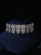 A silver-toned ornate choker necklace with rectangular and teardrop-shaped clear stones, displayed on a dark blue velvet jewelry bust against a black background.