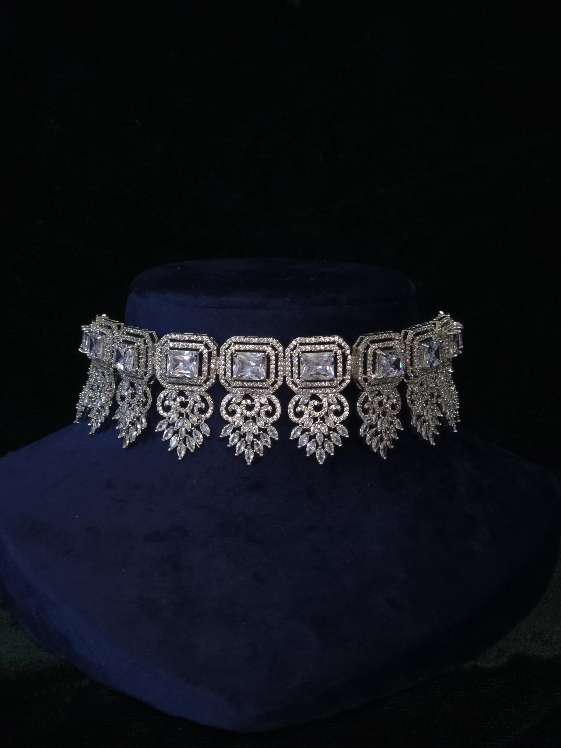 A silver-toned ornate choker necklace with rectangular and teardrop-shaped clear stones, displayed on a dark blue velvet jewelry bust against a black background.