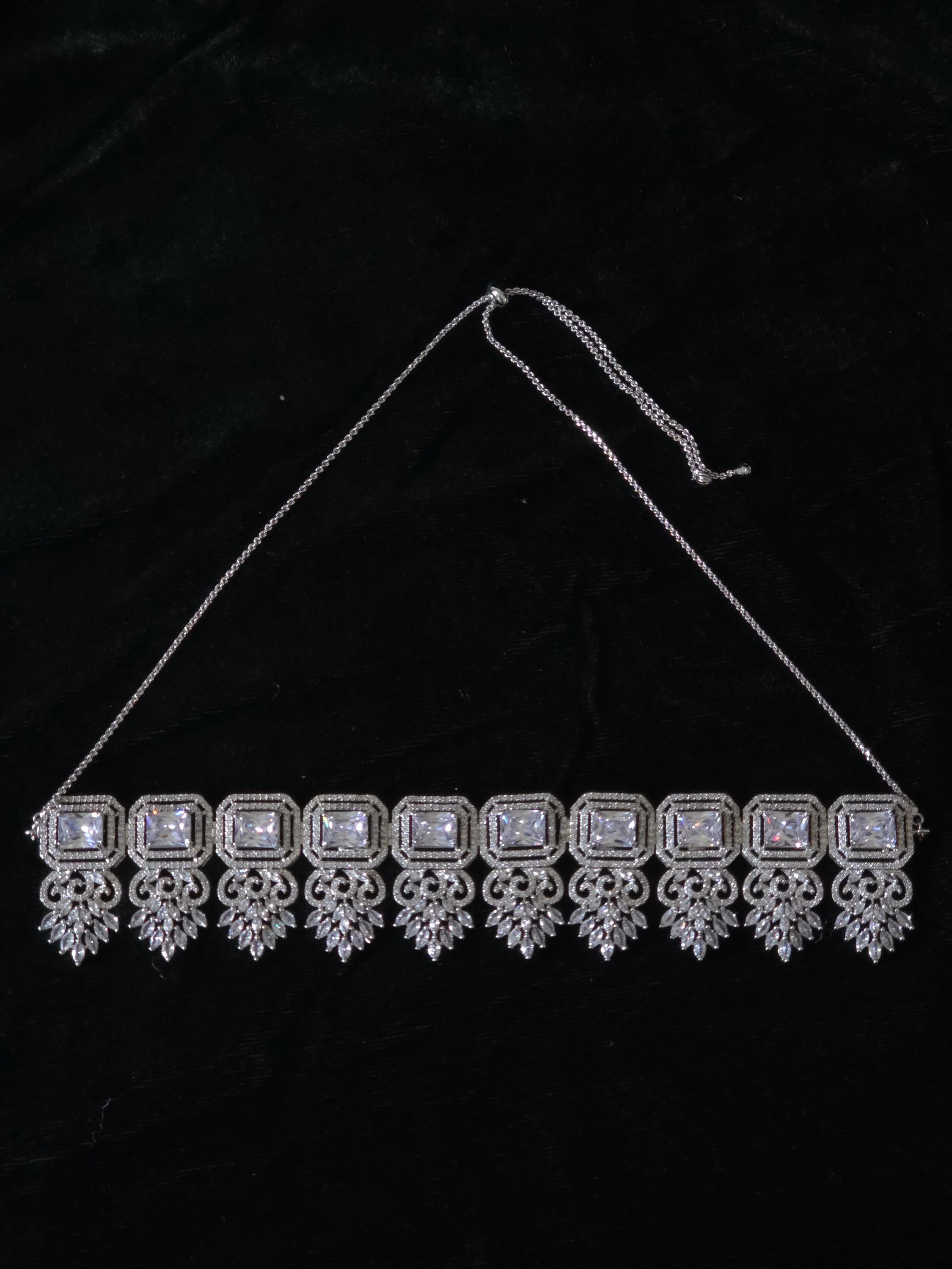 A silver-toned choker necklace with rectangular clear stones and ornate dangling filigree details, displayed against a black fabric background.