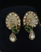 A pair of ornate gold-toned earrings with large oval cream-colored stone centers, surrounded by clear stones and green gemstone floral motifs, with teardrop-shaped clear stones hanging below, displayed on a black background.