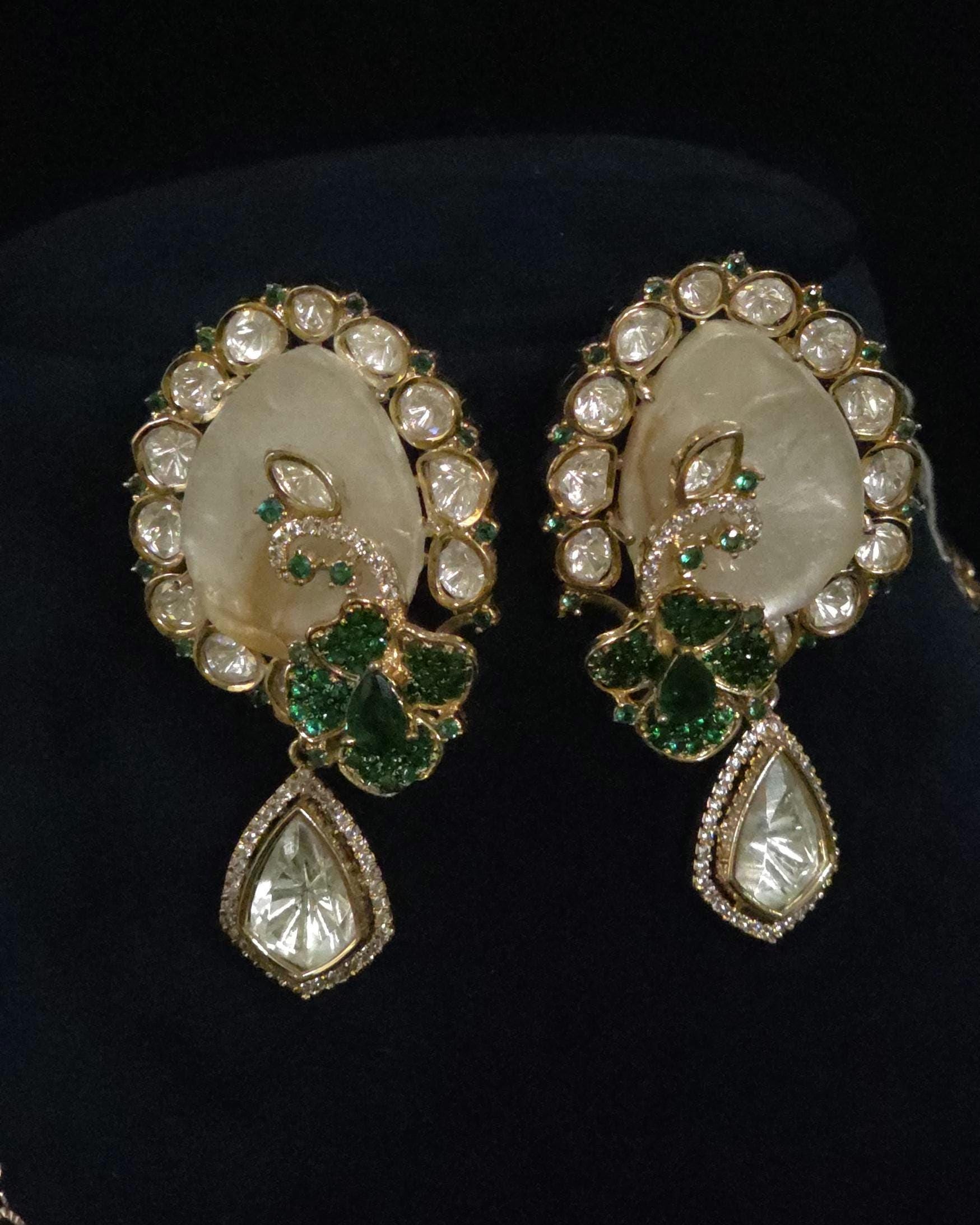 A pair of ornate gold-toned earrings with large oval cream-colored stone centers, surrounded by clear stones and green gemstone floral motifs, with teardrop-shaped clear stones hanging below, displayed on a black background.