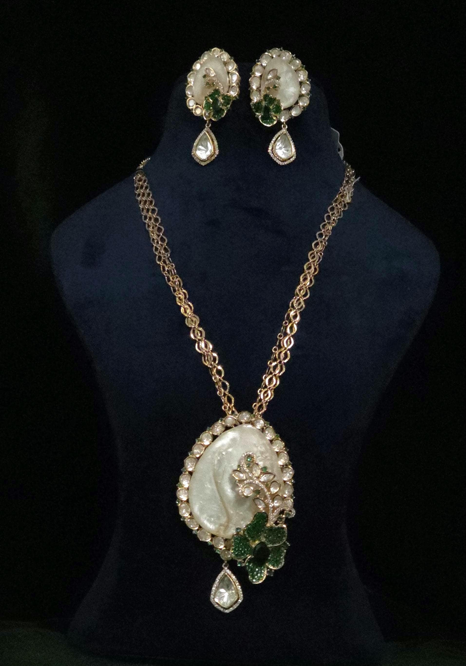 Gold-tone necklace and matching drop earrings displayed on a black velvet jewelry bust. The necklace features a large oval pendant with a white cameo-style face, surrounded by clear rhinestones and a green leaf motif, with a teardrop crystal drop below.