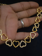 A gold heart-link chain necklace with some hearts paved with clear rhinestones, held in a hand against a dark background.
