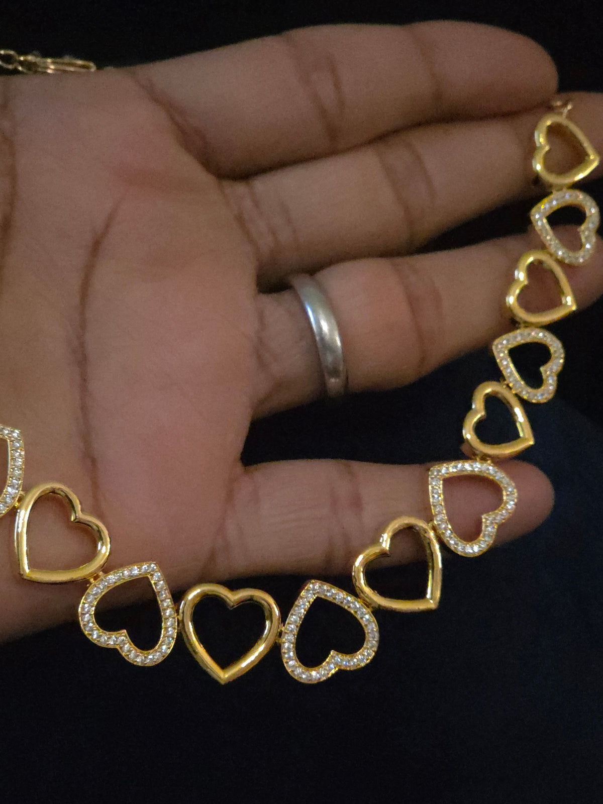 A gold heart-link chain necklace with some hearts paved with clear rhinestones, held in a hand against a dark background.