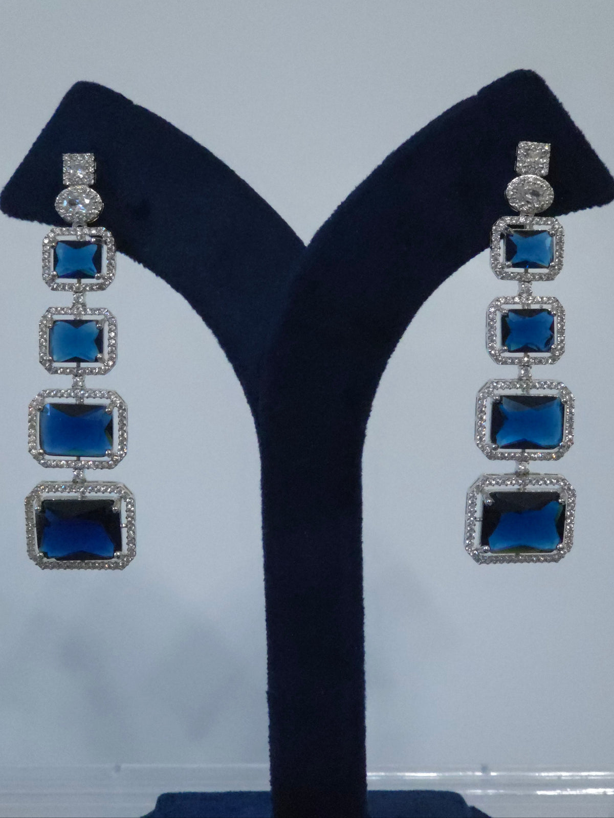 A pair of long drop earrings with four rectangular blue stones in silver settings, displayed on a dark blue velvet jewelry stand 