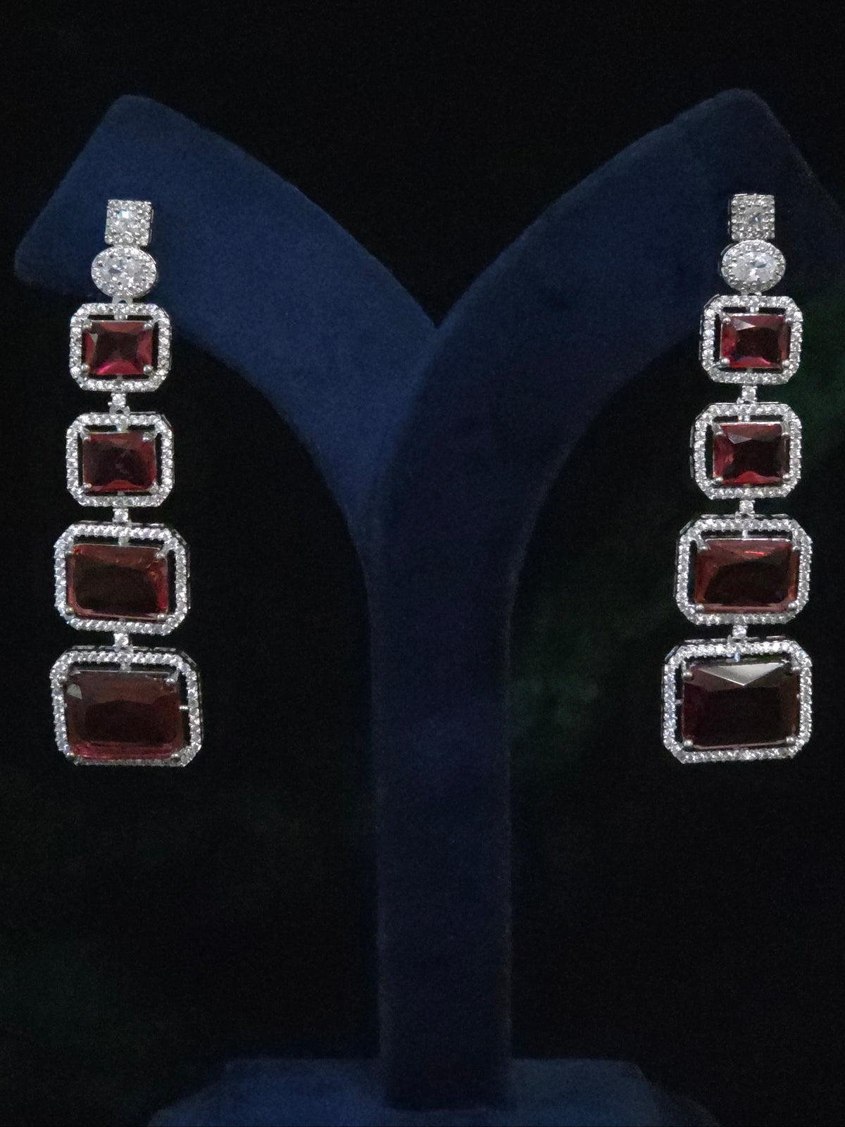 A pair of long drop earrings with four rectangular red stones in silver settings, displayed on a dark blue velvet jewelry stand 