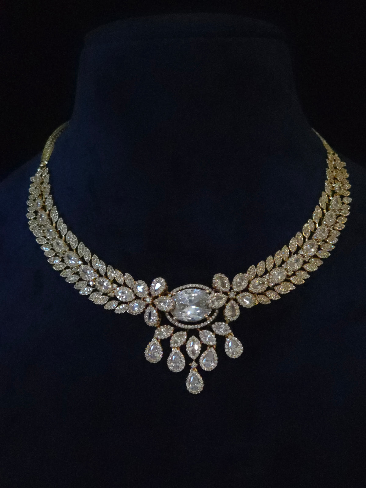 A gold-toned statement necklace with clear teardrop and oval gemstones, displayed on a black velvet jewelry bust.