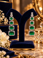 A pair of long drop earrings with four stacked rectangular emerald-cut green gemstones in silver-toned settings surrounded by small clear stones, displayed on a dark blue velvet jewelry stand in an ornate gold-toned room.