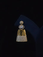 A single statement earring displayed on a black jewelry stand against a black background. The earring features a gold-tone top section with a textured geometric pattern, a round clear gemstone, and a lower section paved with small clear stones.