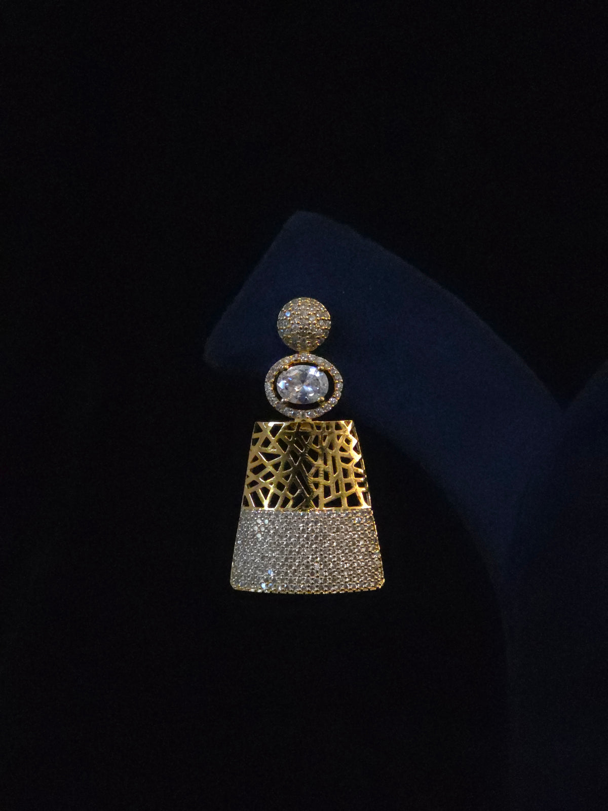 A single statement earring displayed on a black jewelry stand against a black background. The earring features a gold-tone top section with a textured geometric pattern, a round clear gemstone, and a lower section paved with small clear stones.