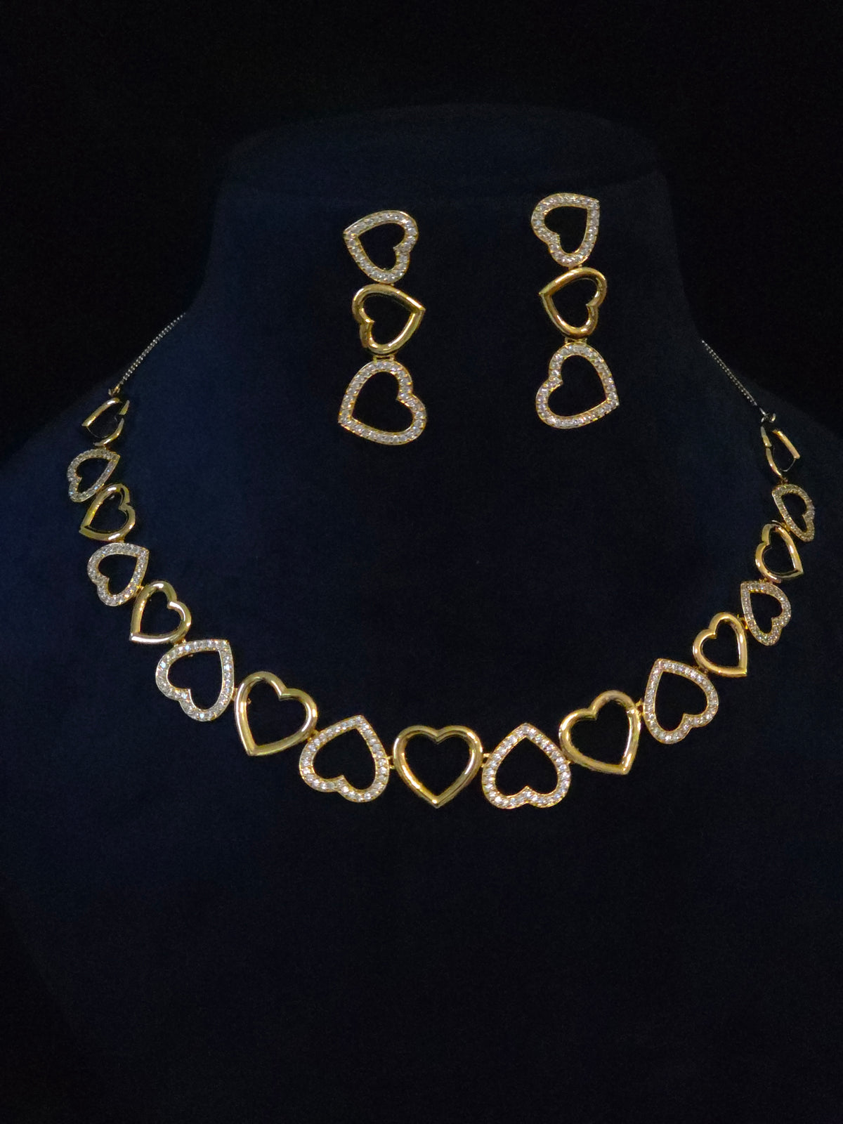 Gold heart link necklace and matching drop earrings displayed on a black velvet jewelry bust. The necklace and earrings feature alternating solid gold and open heart designs, with the open hearts embellished with small clear stones.