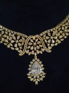 An ornate gold necklace with pavé-set clear stones and a large teardrop-shaped clear gemstone pendant, shown against a dark black background.