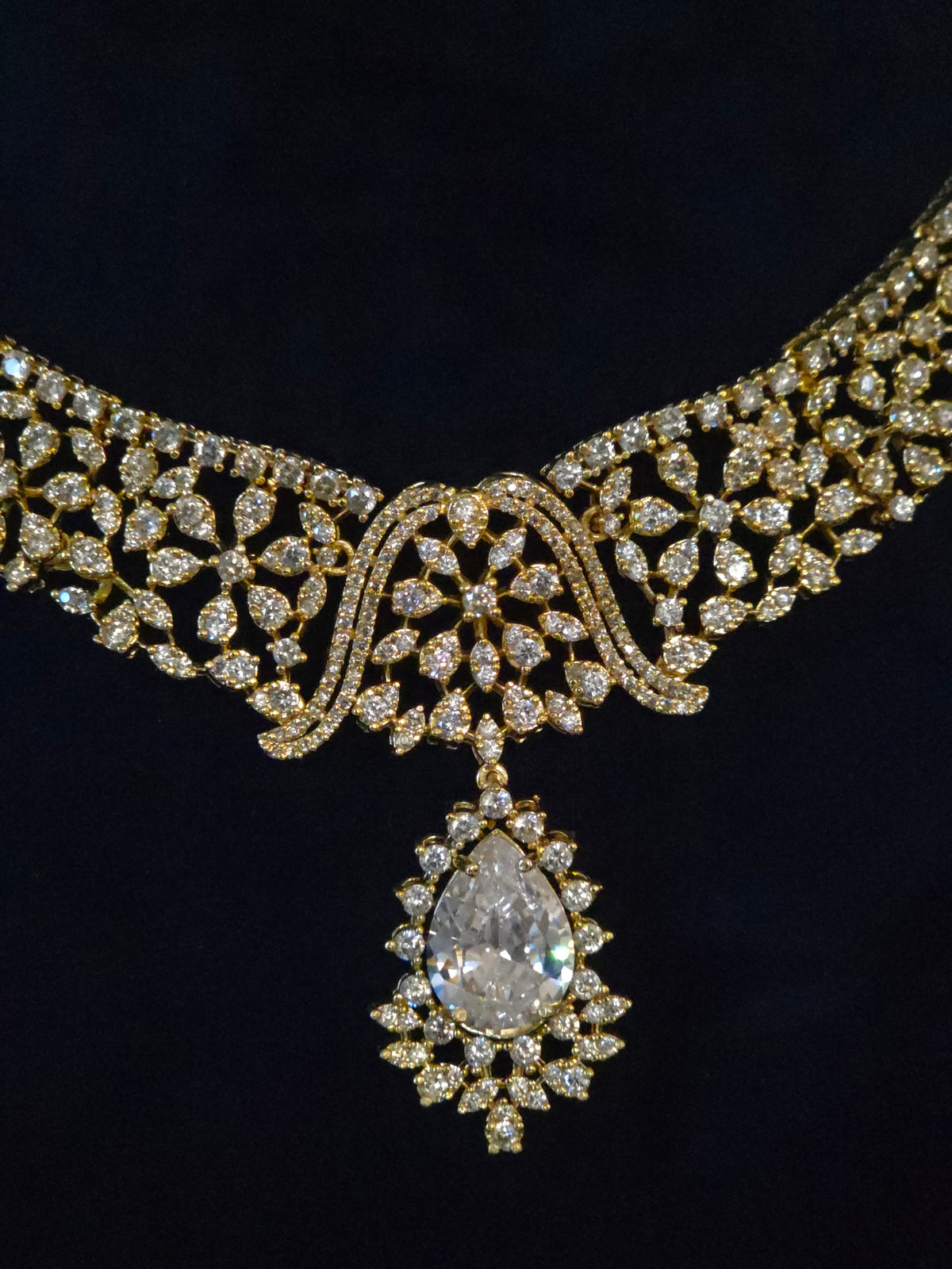 An ornate gold necklace with pavé-set clear stones and a large teardrop-shaped clear gemstone pendant, shown against a dark black background.