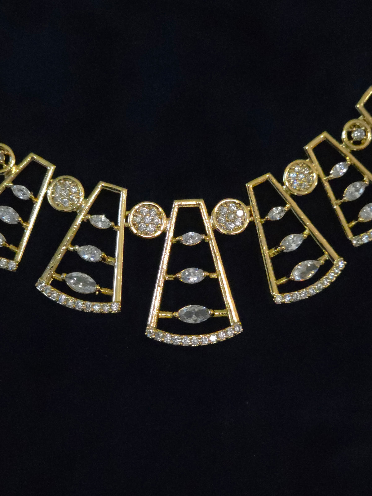 Gold geometric statement necklace with clear marquise and round gemstones, displayed on a black background.