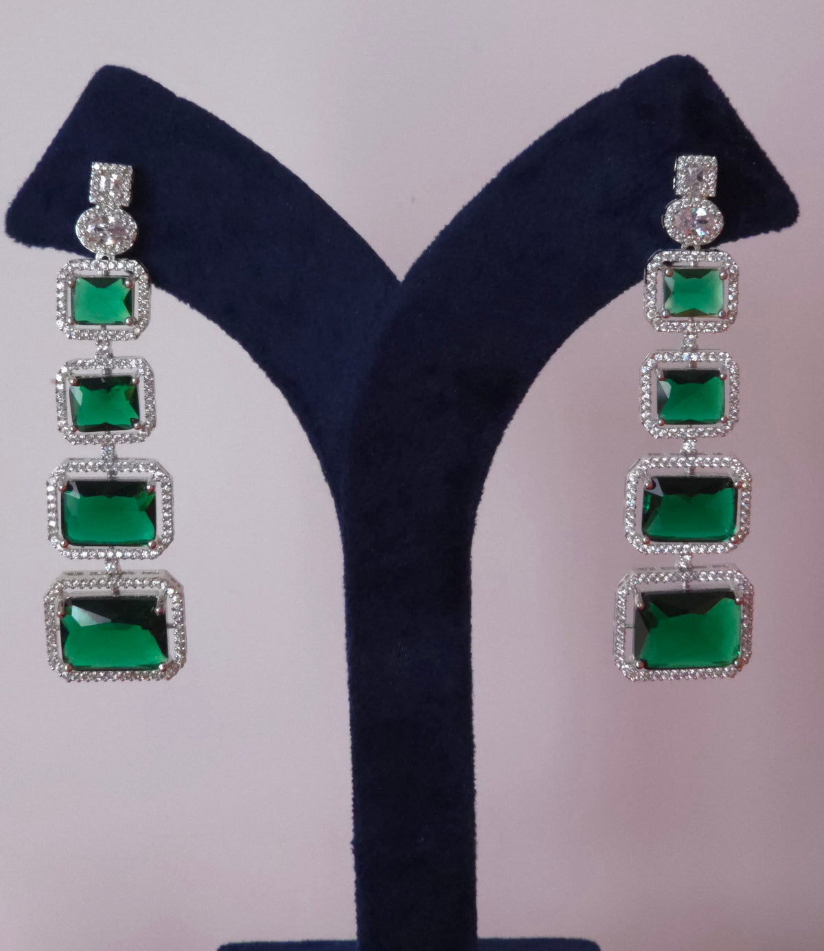 A pair of long drop earrings with five rectangular green stones in silver settings, displayed on a dark blue velvet jewelry stand against a light pink background.