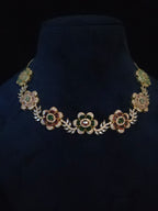 A gold-toned floral necklace with green and red enamel details and clear rhinestone accents, displayed on a dark navy velvet jewelry bust against a black background.