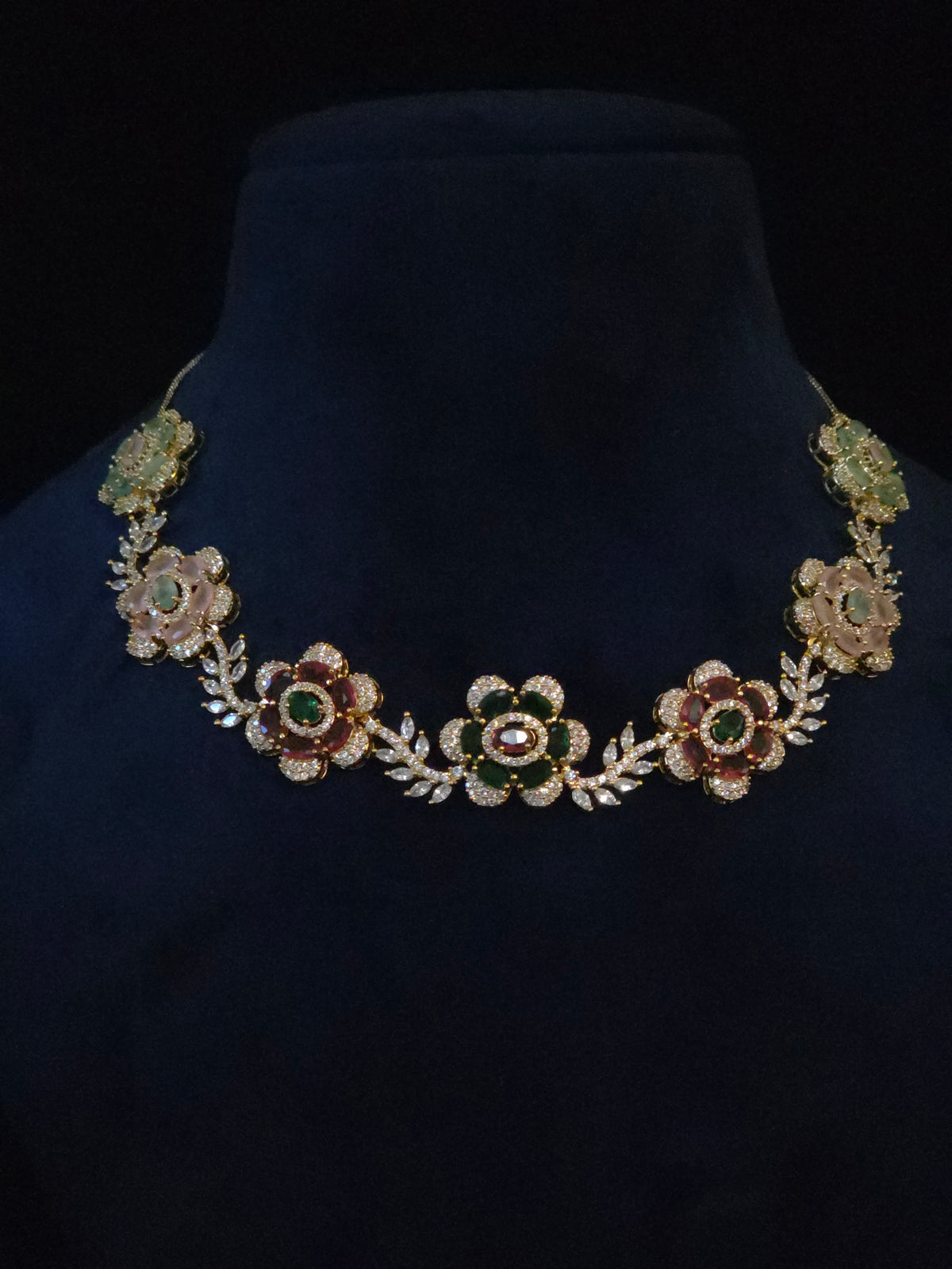 A gold-toned floral necklace with green and red enamel details and clear rhinestone accents, displayed on a dark navy velvet jewelry bust against a black background.