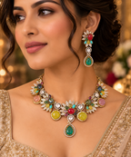 A close-up lifestyle shot of a woman wearing an ornate  jewelry set featuring a multi-colored gemstone necklace and matching drop earrings. The jewelry is embellished with green, red, yellow, pink, and blue stones, worn over a gold embroidered traditional garment.