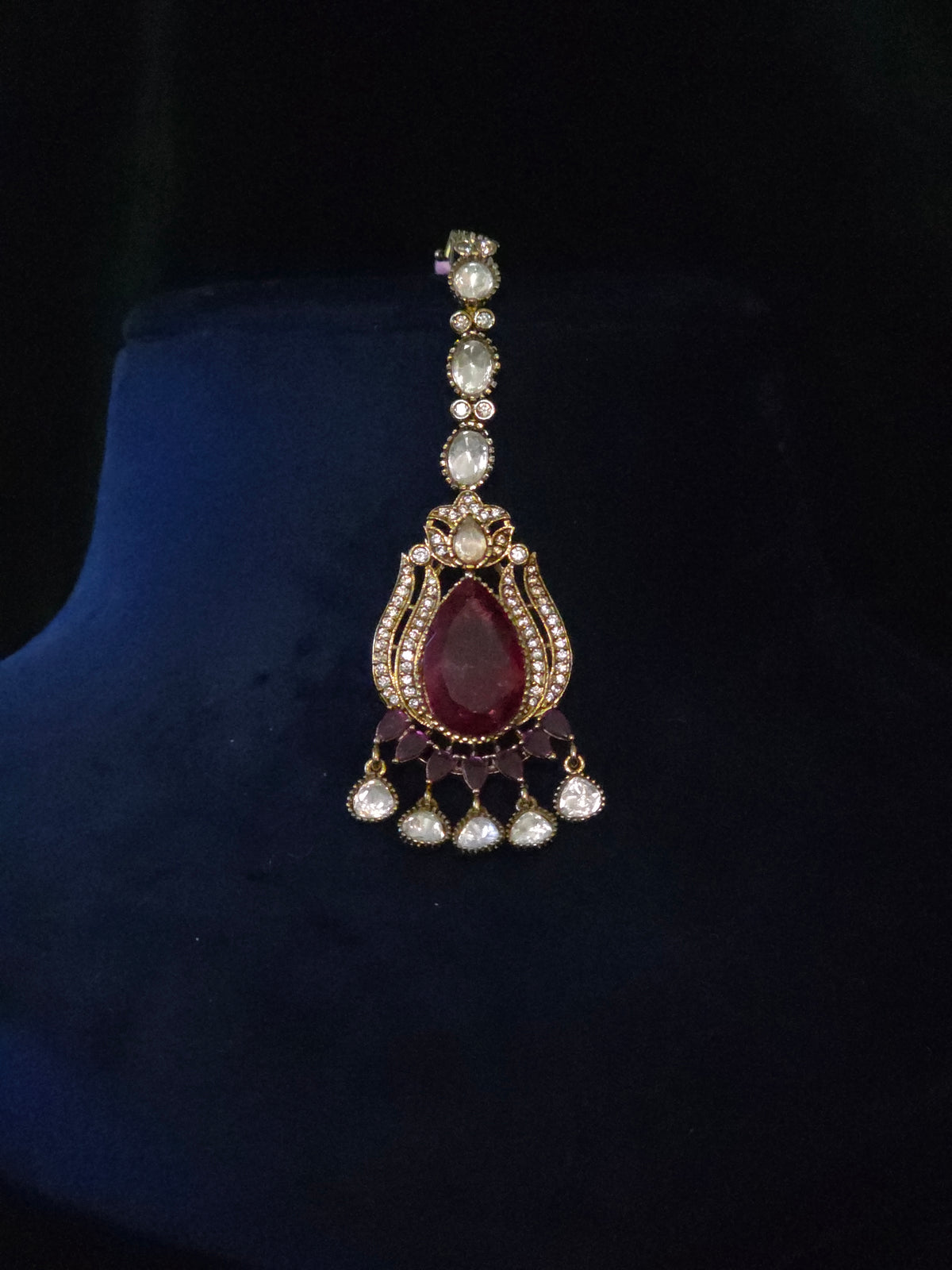 Maang tikka with a deep wine-red center stone, clear accents, and dangling drops on a dark display stand.