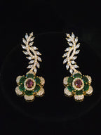 A pair of gold floral drop earrings featuring green emerald-cut stones, a central oval ruby-red stone, and clear pavé-set marquise-cut stones in the leafy top sections, displayed on a black background.