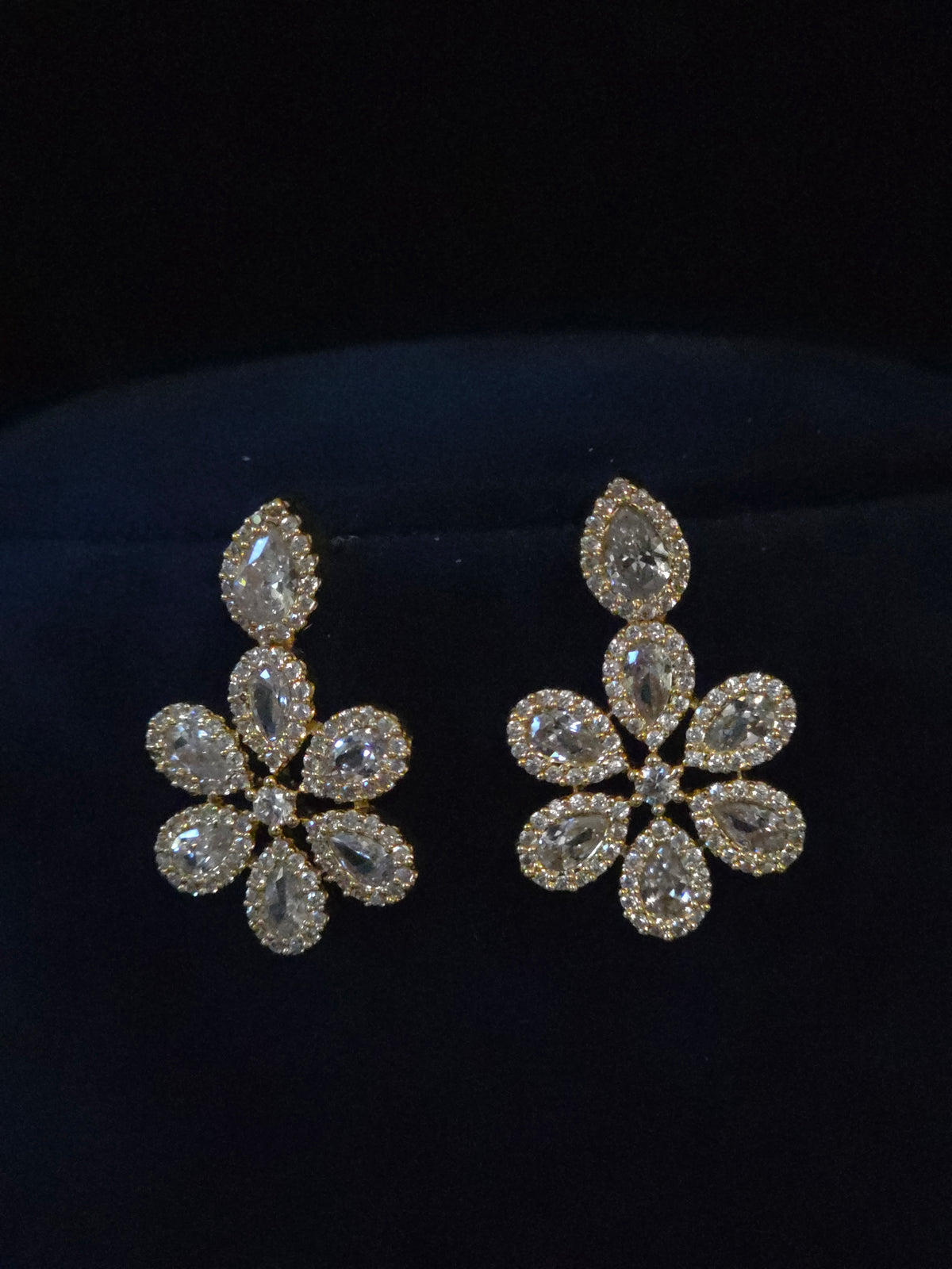 A pair of gold-toned dangle earrings with marquise and round-cut clear crystals arranged in a floral cluster, displayed on a dark velvet surface.