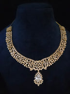 An ornate gold-toned traditional necklace with intricate crystal embellishments and a teardrop-shaped clear central stone, displayed on a dark blue velvet jewelry bust against a black background.