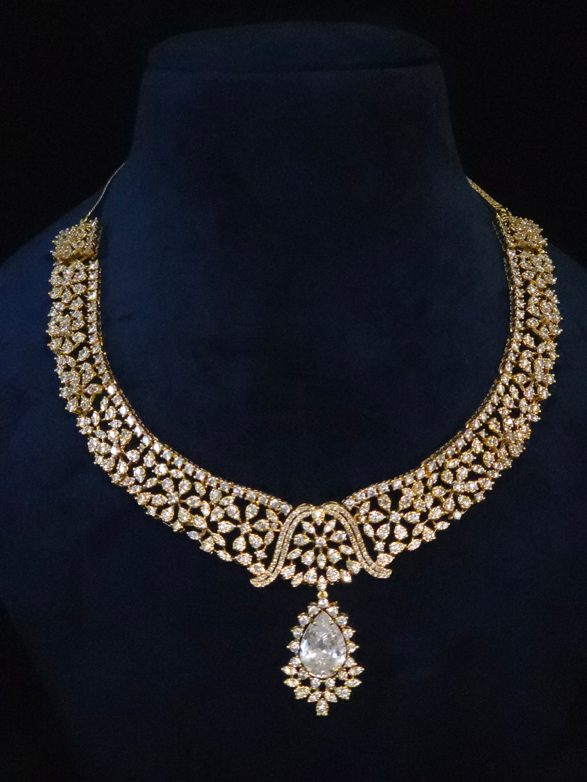 An ornate gold-toned traditional necklace with intricate crystal embellishments and a teardrop-shaped clear central stone, displayed on a dark blue velvet jewelry bust against a black background.