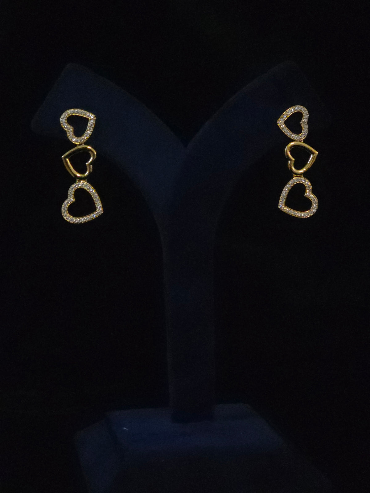 A pair of gold heart drop earrings with small clear stones, displayed on a black velvet jewelry stand against a black background.