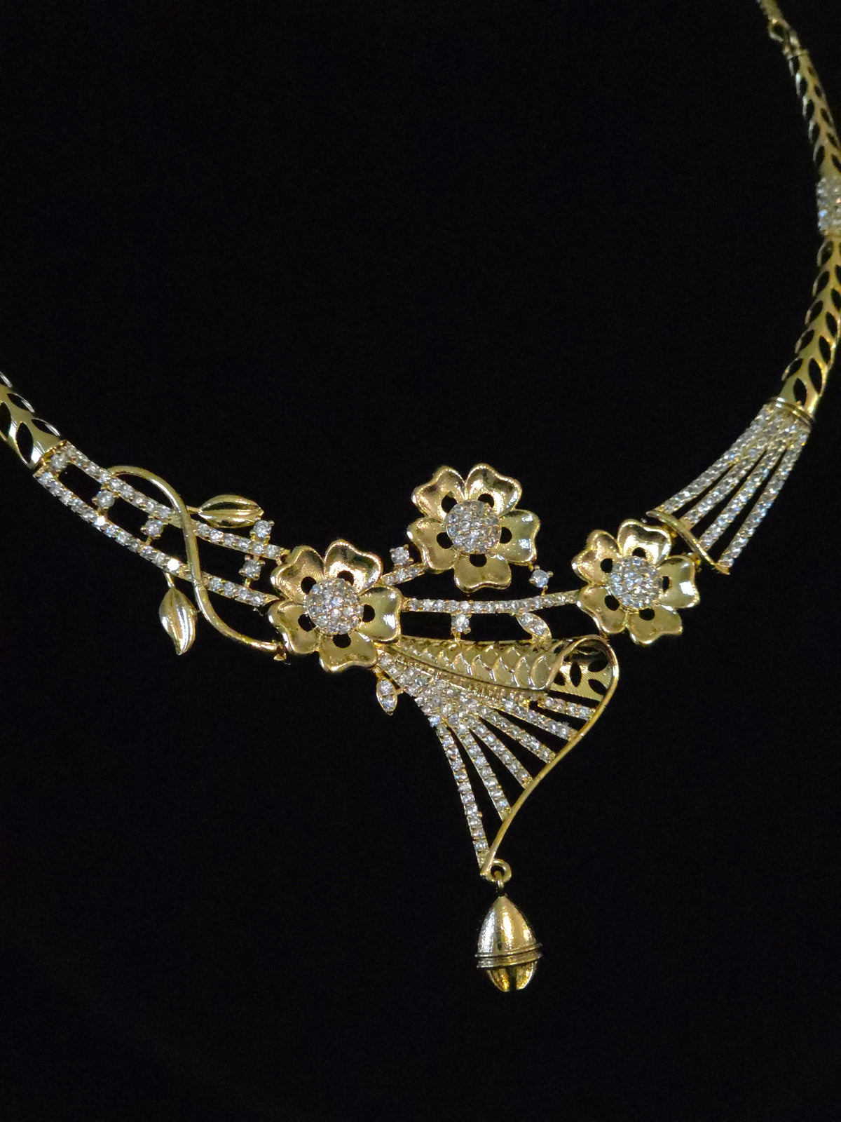 A gold-toned Art Deco style necklace featuring floral motifs, pavé-set clear stones, and a dangling bell-shaped pendant, displayed against a solid black background.
