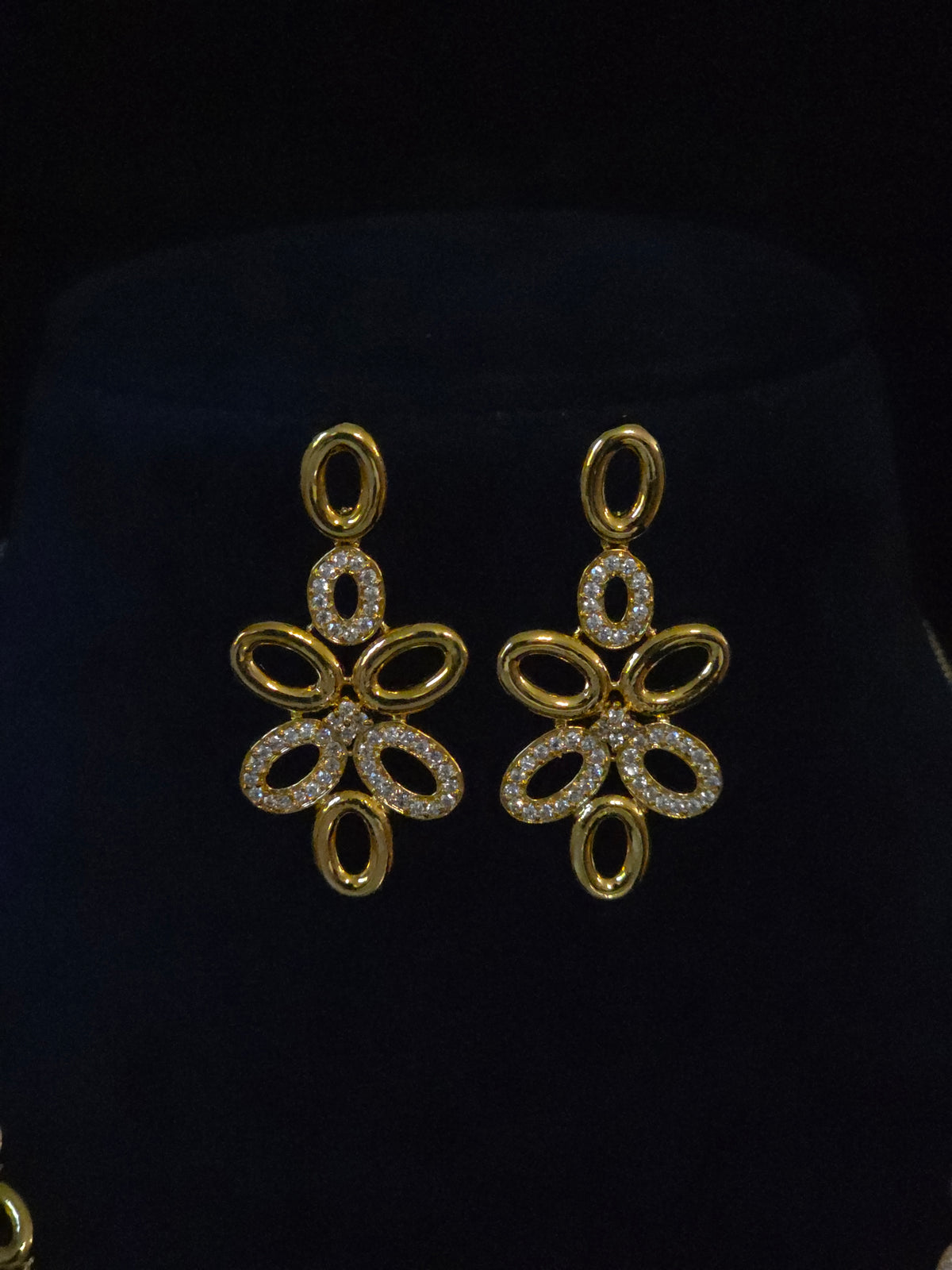A pair of gold-toned dangle earrings with openwork floral or butterfly-like cutouts, each adorned with small clear stones, displayed on a black velvet jewelry bust.
