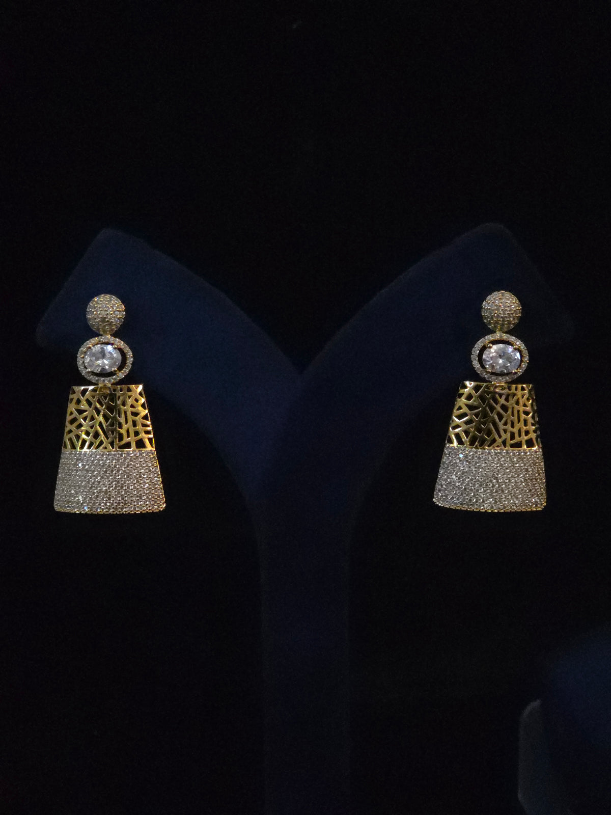 A pair of gold and silver-toned statement earrings displayed on a black velvet jewelry stand. Each earring features a round crystal-studded top, a textured gold middle section, and a silver crystal-encrusted lower section.