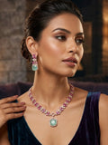 A woman wearing a pink gemstone necklace with a mint green pendant and matching drop earrings, styled with a dark purple velvet dress in a dimly lit interior setting.