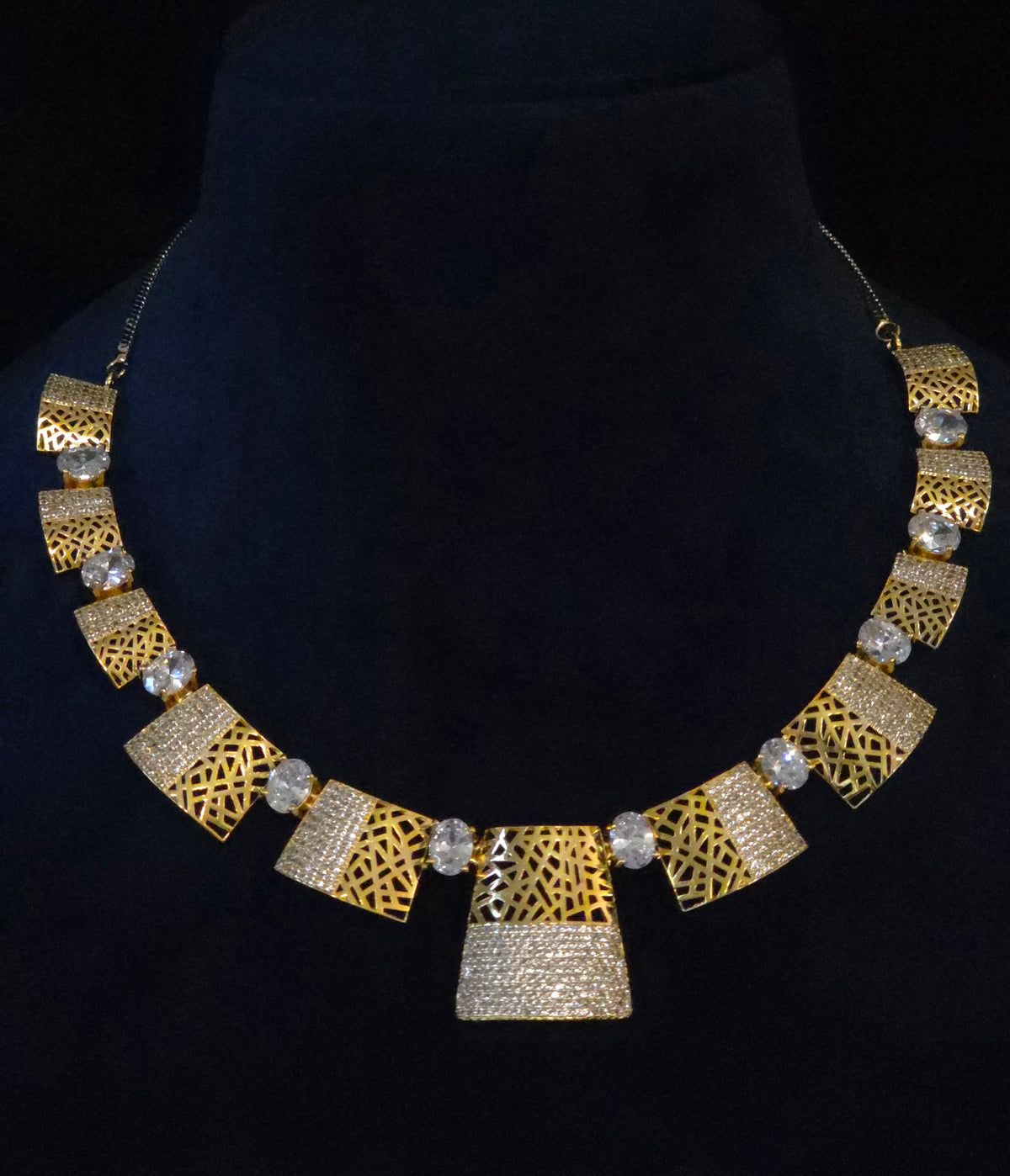 A statement bib necklace displayed on a black velvet jewelry bust. The necklace features alternating sections of textured gold geometric filigree, textured silver geometric panels, and small round clear crystal accents.