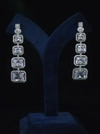 A pair of long drop earrings with four rectangular clear stones in silver settings, displayed on a dark blue velvet jewelry stand 