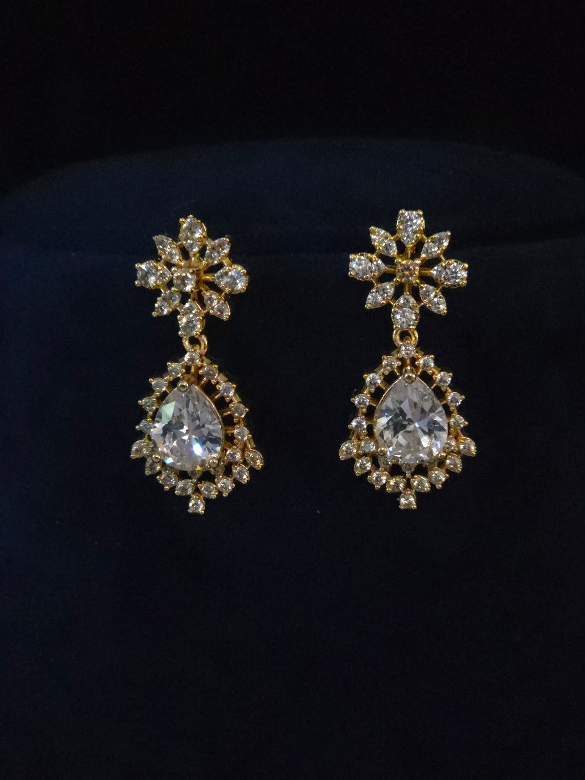 A pair of gold-toned drop earrings with clear teardrop-shaped crystals and surrounding clear stones, displayed on a black velvet surface.