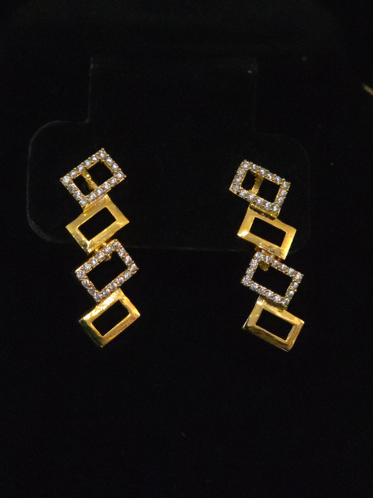 A pair of gold geometric drop earrings with rectangular links, some bordered in clear stones, displayed on a black background.