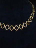 Gold geometric open-link necklace with diamond-shaped links, some set with clear stones, displayed on a dark navy background.