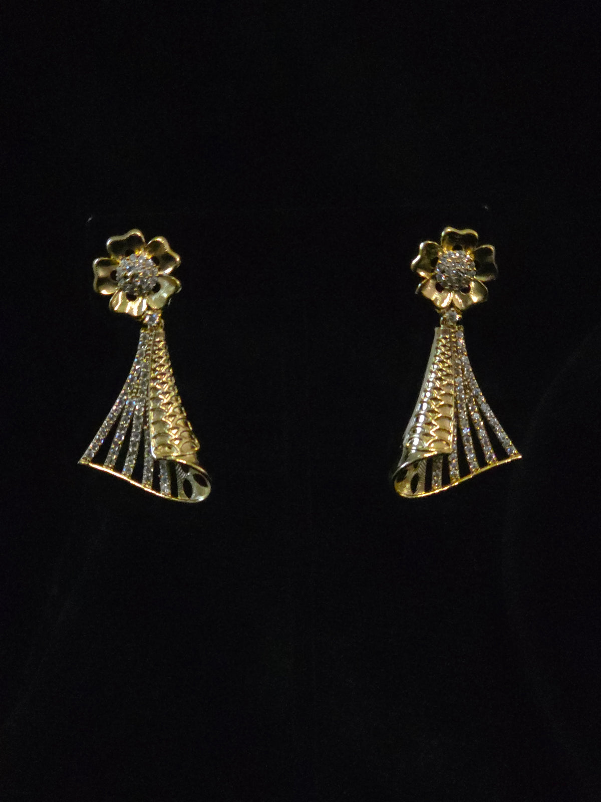 A pair of gold-toned statement earrings with a flower-shaped top set with clear stones and a textured, fan-like drop below, displayed against a solid black background.