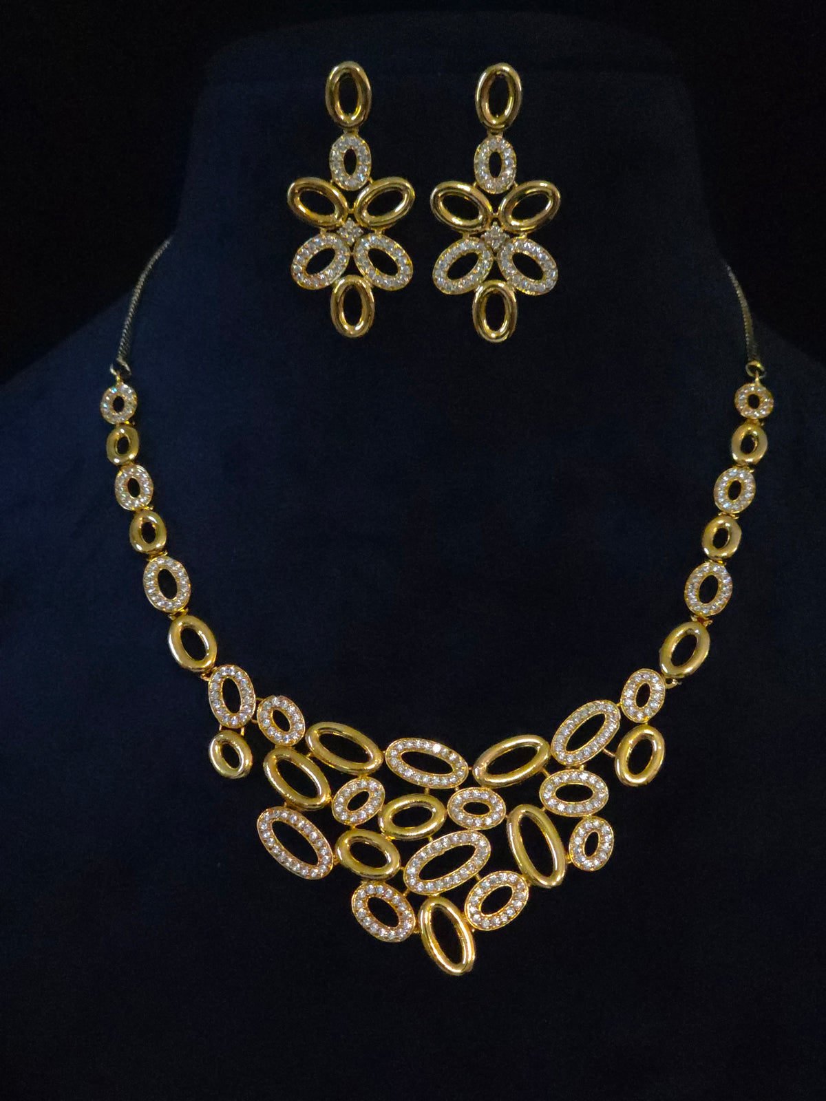 Gold-tone jewelry set featuring a statement collar necklace and matching drop earrings, both adorned with clear rhinestones, displayed on a black velvet bust.