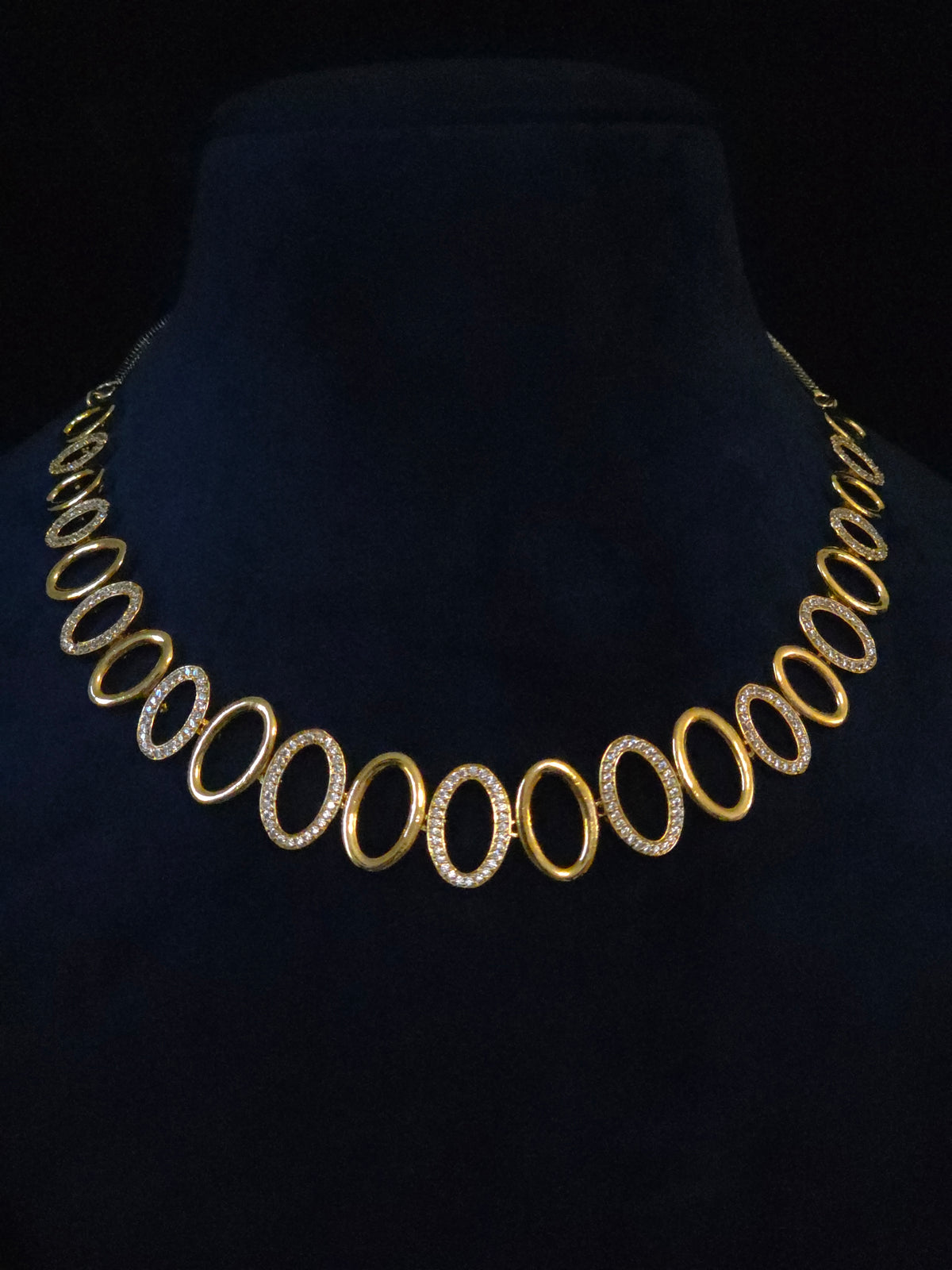 A gold oval-link necklace with alternating links set with small clear stones, displayed on a black velvet jewelry bust.