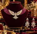 A luxury jewelry set displayed on a dark burgundy velvet mannequin bust, featuring a winged necklace with pink gemstones and crystal-encrusted gold details, matching drop earrings, and a small T-shaped stand, set against a richly decorated background with gold frames and floral arrangements.