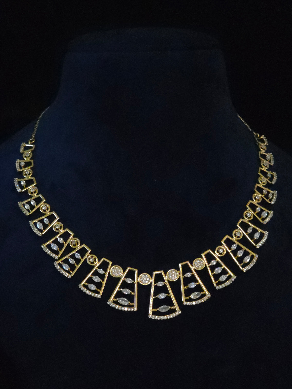 A gold statement necklace with geometric fan-shaped motifs and small clear stones, displayed on a black velvet jewelry bust against a black background.